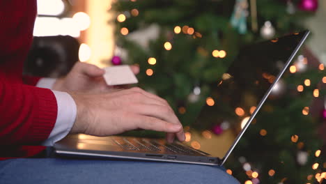 Close-Up-View-Of-Man-Hands-Typing-On-Laptop-Over-Her-Legs-Sitting-Near-The-Christmas-Tree-In-Living-Room-With-Christmas-Decoration