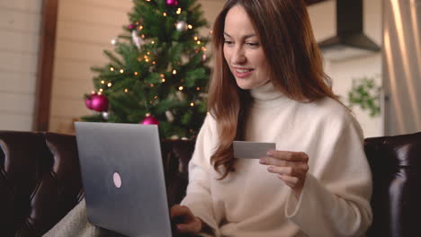 Glückliche-Frau,-Die-Online-Mit-Kreditkarte-Einkauft,-Während-Sie-Zu-Hause-Auf-Dem-Sofa-Neben-Einem-Weihnachtsbaum-Im-Wohnzimmer-Sitzt