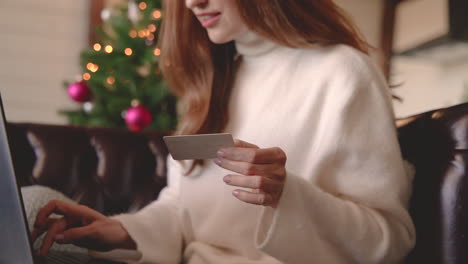 Nahaufnahme-Von-Frauenhänden,-Die-Online-Mit-Kreditkarte-Einkaufen,-Während-Sie-Zu-Hause-Auf-Dem-Sofa-Neben-Einem-Weihnachtsbaum-Im-Wohnzimmer-Sitzen