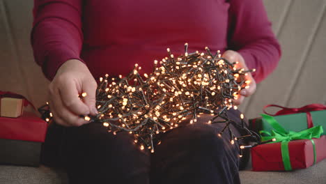 Close-Up-View-Of-A-Woman's-Hands-Holding-Christmas-Lights-Sitting-On-The-Sofa-With-Christmas-Gifts-Around