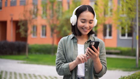 Happy-Young-Japanese-Girl-Listening-To-Music-In-Wireless-Headphones-While-Walking-Outdoors