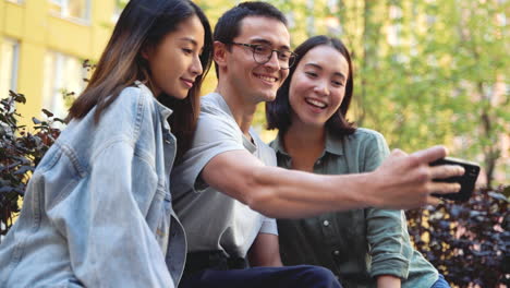 Young-Japanese-Man-Holding-Mobile-Phone-And-Taking-A-Funny-Selfie-Photo-With-His-Two-Female-Friends-While-Sitting-Outdoors-In-The-Park