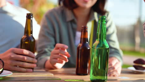 Close-Up-Of-Three-Unrecognizable-Friends-Talking-And-Drinking-Beer-While-Sitting-At-Table-Outdoors-In-A-Sunny-Day