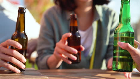 Close-Up-Of-Three-Unrecognizable-Friends-Toasting-With-Beer-While-Sitting-At-Table-Outdoors-In-A-Sunny-Day