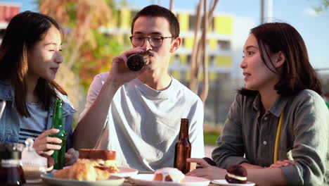 Gruppe-Von-Drei-Japanischen-Freunden,-Die-Sich-Unterhalten-Und-Bier-Trinken,-Während-Sie-An-Einem-Sonnigen-Tag-Im-Freien-Am-Tisch-Sitzen-2