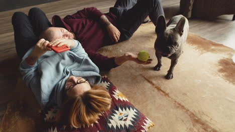 Top-View-Of-Couple-Playing-With-Her-Bulldog-Dog-With-A-Tennis-Ball-Liying-On-The-Floor-In-Living-Room
