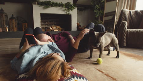 Couple-Playing-With-Her-Bulldog-Dog-With-A-Tennis-Ball-Liying-On-The-Floor-In-Living-Room