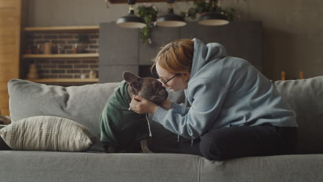 Red-Haired-Woman-Puts-A-Hood-On-Her-Bulldog-Dog-Head-And-Caresses-Her-While-Sitting-On-The-Couch-In-The-Living-Room-At-Home