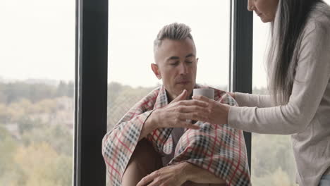 Sick-Middle-Aged-Man-With-Blanket-On-His-Shoulder-Sitting-At-Window-While-His-Woman-Giving-Him-A-Hot-Drink-And-Taking-Care-Of-Him