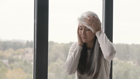 Sick-Woman-Having-Headache-And-Holding-An-Ice-Pack-To-Her-Head-While-Sitting-By-The-Window-At-Home