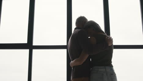 Back-View-Of-Middle-Aged-Man-Embracing-His-Sad-Wife-And-Trying-To-Comfort-Her-While-Standing-Together-At-Window-At-Home-1