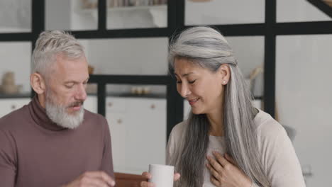 Middle-Aged-Woman-Having-Chest-Pain-And-Her-Husband-Giving-Her-A-Hot-Drink-While-They-Sitting-Together-On-Sofa-At-Home