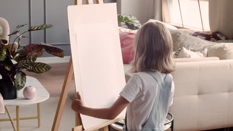 Rear-View-Of-Little-Blonde-Girl-Painting-On-The-Lectern-Next-To-The-Window-At-Home