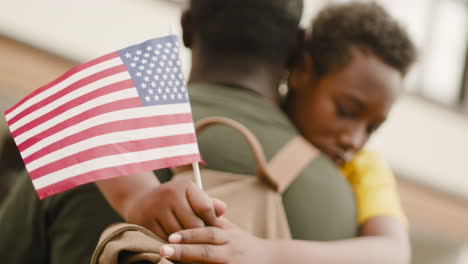 Portrait-Of-A-Sad-Boy-Holding-An-Usa-Flag-And-Embracing-His-Unrecognizable-Military-Father