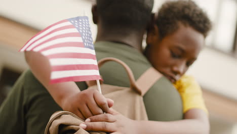 Portrait-Of-A-Sad-Boy-Holding-An-Usa-Flag-And-Embracing-His-Unrecognizable-Military-Father-1