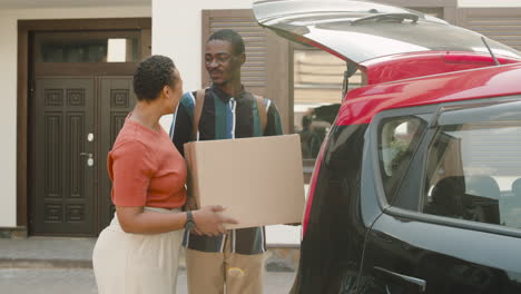 Young-Man-Holding-Carton-Box-And-Putting-It-In-Car-Trunk-While-Talking-With-His-Mom-Standing-Next-To-Him