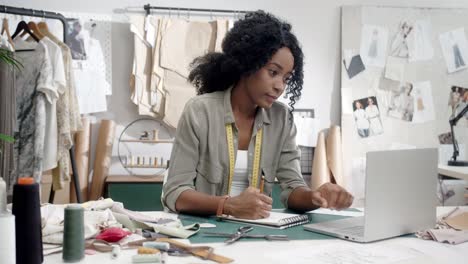 Woman-Clothes-Designer-Sitting-And-Working-At-The-Laptop-Computer,-Drawing-Outlines-In-Her-Notebook-And-Smiling-To-The-Camera-In-Her-Studio