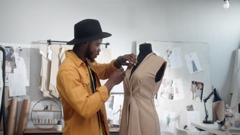 Stylish-Young-Man-Clothing-Designer-Wearing-Hat-Making-Marks-With-Pins-On-Dress-In-Mannequin-In-The-Studio