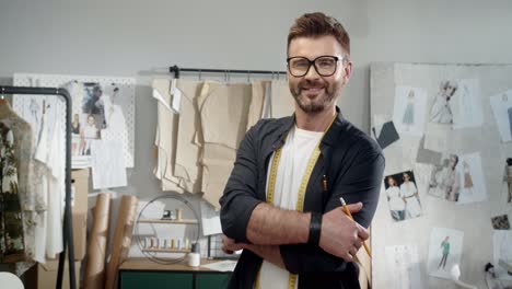 Young-Male-Designer-In-Eyeglasses-In-The-Workshop-Measure-Pieces-Of-Clothes-In-Studio-And-Smiles-At-Camera
