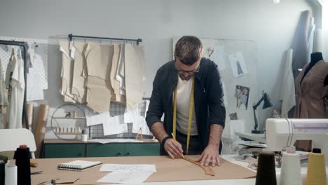 Young-Male-Designer-In-Eyeglasses-In-The-Workshop-Measure-Pieces-Of-Clothes-In-Studio