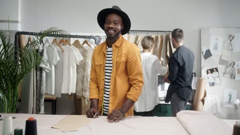 Young-Designer-Takes-Measurements-And-Smiles-At-Camera-While-A-Man-And-Blonde-Woman-Colleagues-Choose-Clothes-On-The-Background