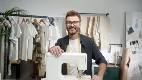 Man-Tailor-In-Glasses-Sitting-At-The-Table-With-A-Sewing-Machine-In-His-Workshop-And-Smiling-Cheerfully-To-The-Camera