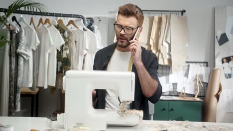 Man-Tailor-In-Glasses-Sitting-At-The-Table-With-A-Sewing-Machine-And-Choosing-Clothes-Pieces-And-Talking-On-The-Smartphone