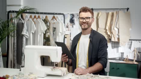 Man-Tailor-In-Glasses-Sitting-At-The-Table-With-A-Sewing-Machine-And-Using-Tablet,-Then-Smiles-At-Camera