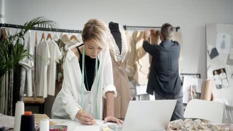 Woman-Tailor-Taking-Notes-On-The-Table,-Then-Colleage-Man-Aproachs-Her-And-They-Choose-Clothes-Pieces