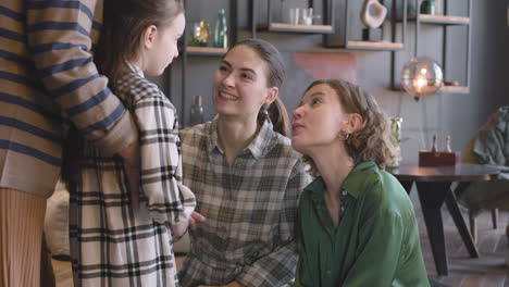 Little-Girl-Talking-With-Her-Mother-And-Aunt-While-Unrecognizable-Grandmother-Hugging-Her-From-Behind-During-A-Family-Reunion-At-Home