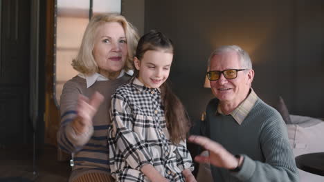 Glückliche-Großeltern-Mit-Ihrer-Kleinen-Enkelin-Lächeln-Und-Winken-In-Einem-Modernen-Wohnzimmer-Zu-Hause-In-Die-Kamera