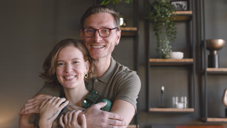 Portrait-Of-A-Happy-Couple-Smiling-At-Camera-At-Home-While-Man-Embracing-Woman-From-Behind-1