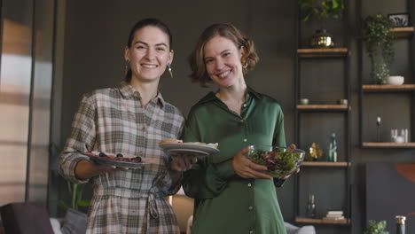 Zwei-Glückliche-Frauen-Stehen-Mit-Essen-Im-Wohnzimmer-Und-Schauen-In-Die-Kamera