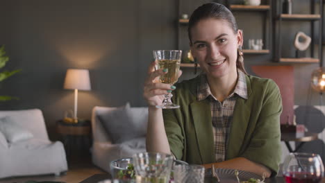 Portrait-Of-A-Smiling-Pretty-Woman-Sitting-At-Dinner-Table-And-Toasting-To-Camera-With-White-Wine-Glass