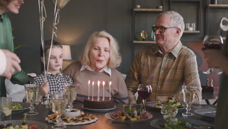 Seniorin-Bläst-Kerzen-Auf-Geburtstagstorte-Während-Einer-Feier-Mit-Ihrer-Familie-Zu-Hause-Aus