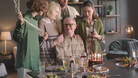 Senior-Mann-Bläst-Kerzen-Auf-Geburtstagstorte-Während-Einer-Feier-Mit-Seiner-Familie-Zu-Hause-Aus