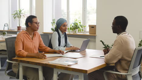 Una-Mujer-Y-Una-Compañera-De-Trabajo-Musulmana-Entrevistan-A-Un-Joven-Sentado-En-Una-Mesa-En-La-Oficina-5