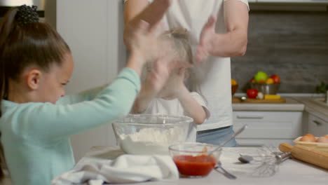 Medium-Shot-Of-Two-Adorable-Little-Girls-Of-Elementary-School-Age-And-Their-Father-Cooking-Something-Together-And-Clapping-Hands-To-Clean-Them-From-Flour