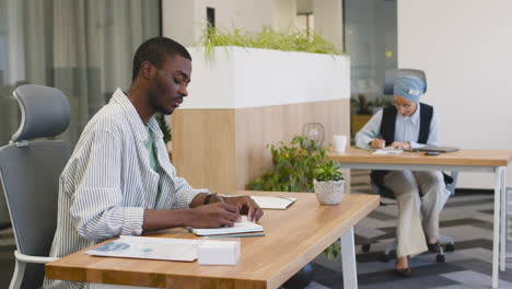 Young-Smiling-Worker-Working-Taking-Notes-Sitting-At-His-Desk