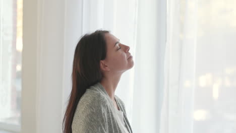 Woman-With-Closed-Eyes-Deeply-Breathing-While-Standing-Near-The-Window-At-Home-In-The-Morning
