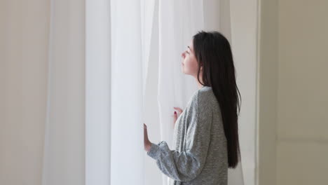 Pensive-Woman-Standing-Looking-Outside-Through-A-Window-At-Home-In-The-Morning