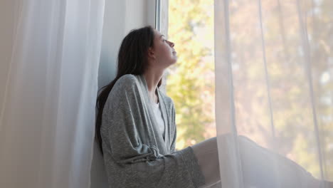 Pensive-Woman-Sitting-On-A-Windowsill,-Looking-Outside-Through-The-Window-In-The-Morning-At-Home