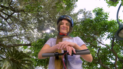 View-From-Below-Of-A-Funny-Young-Woman-With-Electric-Scooter-And-Wearing-Helmet-Showing-Tongue-At-Camera