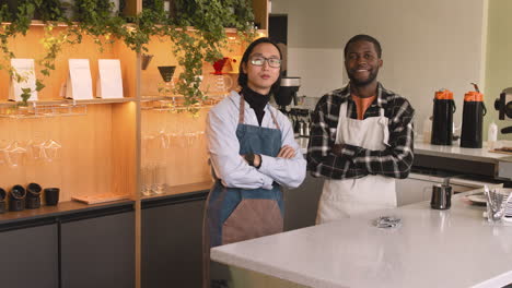 Two-Multiethnic-Waiters-With-Crossed-Arms-Smiling-At-Camera-While-Standing-Behind-Counter-In-A-Coffee-Shop