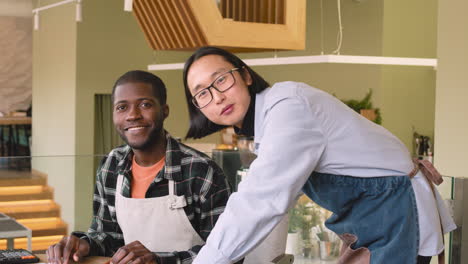 Two-Multiethnic-Coffee-Shop-Owners-Smiling-At-Camera-While-Calculating-Finance-Bills-On-Laptop-Computer