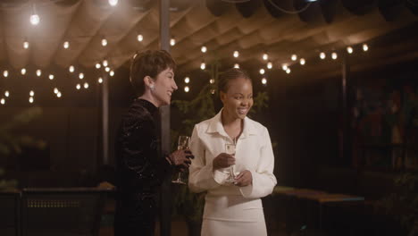 Two-Elegant-Multiethnic-Women-With-Champagne-Glass-Talking-To-Each-Other-And-Then-Welcoming-Two-Men-To-New-Year's-Eve-Party