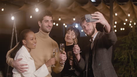 Group-Of-Four-Happy-Multiethnic-Friends-Taking-A-Selfie-Video-And-Toasting-With-Champagne-Glasses-At-New-Year's-Eve-Party