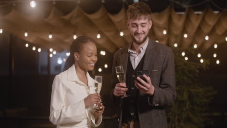 Man-And-Woman-Toasting-With-Champagne-Glasses-While-Taking-A-Selfie-Video-At-New-Year's-Eve-Party