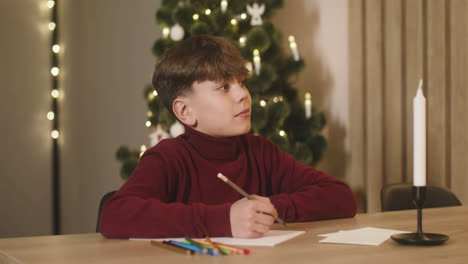 Junge-Im-Roten-Rollkragenpullover,-Der-Einen-Wunschbrief-Schreibt,-Der-An-Einem-Tisch-In-Einem-Mit-Einem-Weihnachtsbaum-Geschmückten-Raum-Sitzt