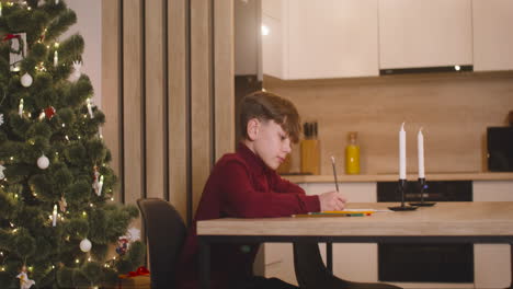 Side-View-Of-A-Boy-In-Red-Turtleneck-Writing-A-Letter-Of-Wishes-Sitting-At-A-Table-In-A-Room-Decorated-With-A-Christmas-Tree